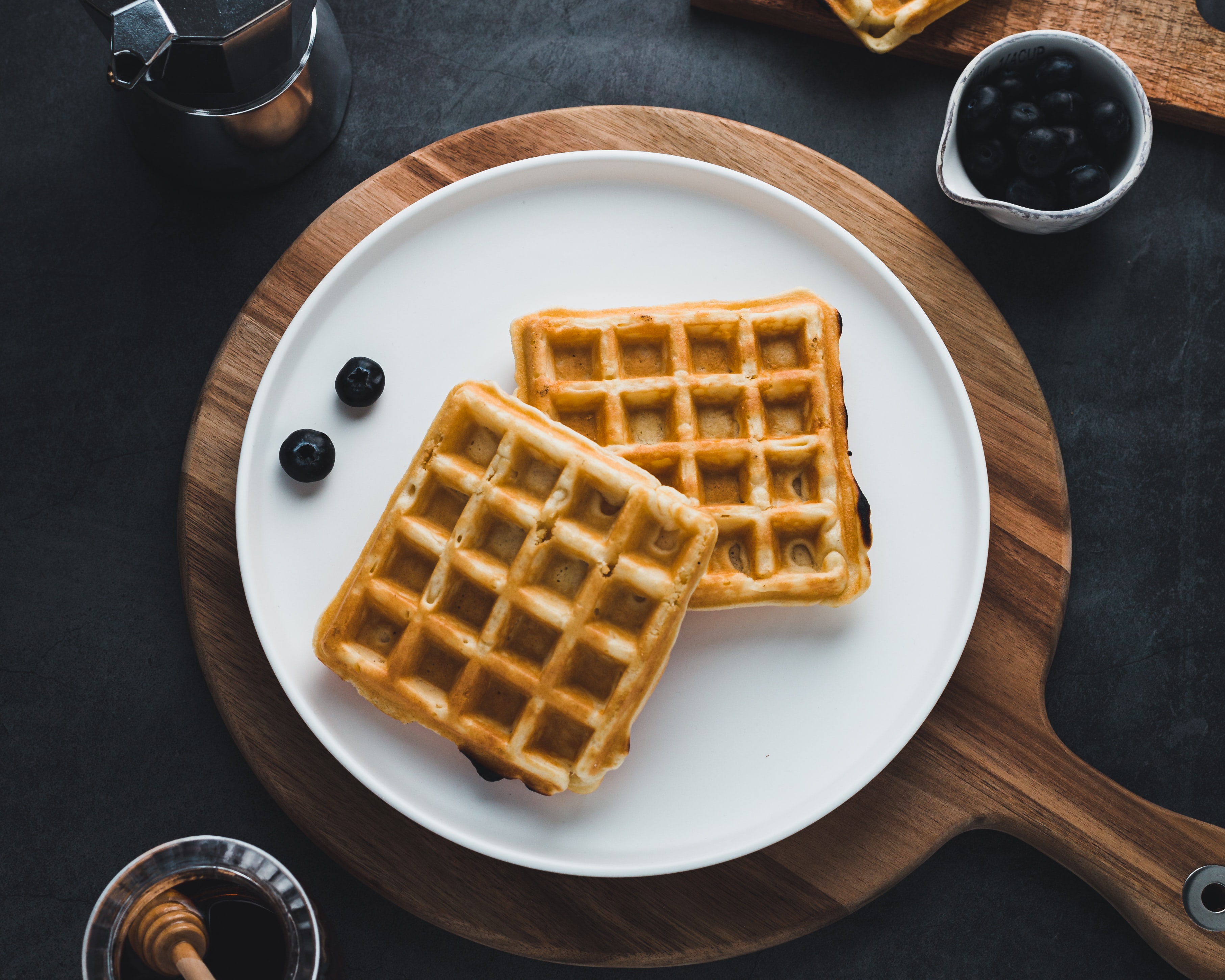 waffle em prato branco em cima de uma tabua de madeira acompanhado de mirtilo e mel 